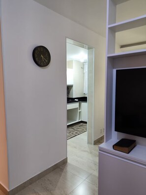 Un pasillo con un reloj, una zona de cocina con armarios y un televisor sobre un mueble.