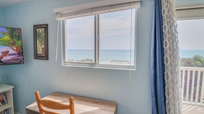 A room with a view of the ocean, a wooden chair, a small table, a framed picture, and a bookshelf. - A-Frame in the Sand - Oceanfront + Pet-Friendly (New Smyrna Beach)