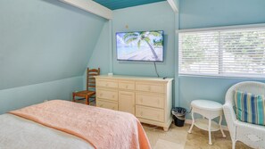 A bedroom with a bed, dresser, chair, and a TV displaying a beach scene. - A-Frame in the Sand - Oceanfront + Pet-Friendly (New Smyrna Beach)