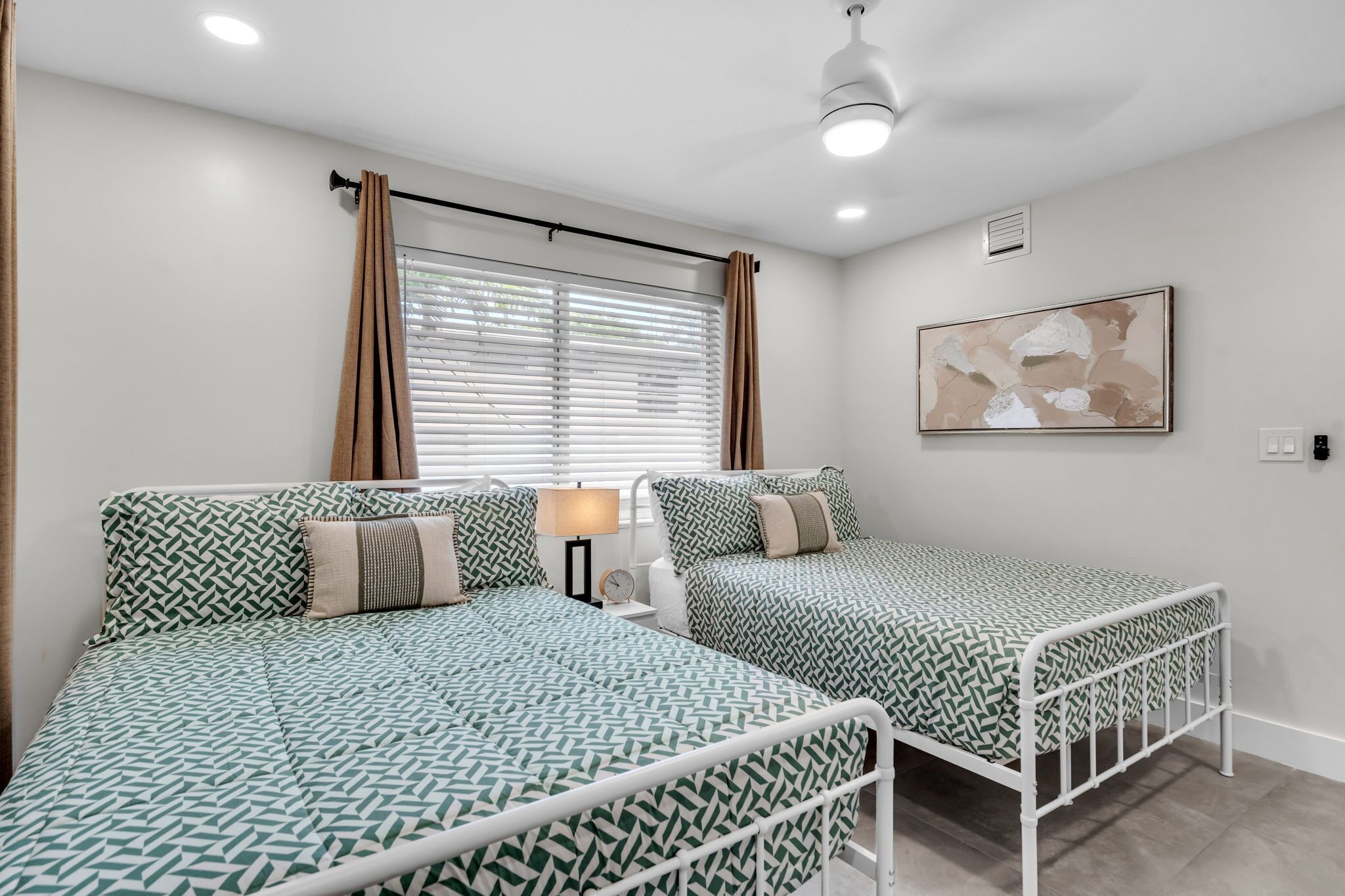 A room with two beds, a nightstand, a lamp, a ceiling fan, and a window with curtains.