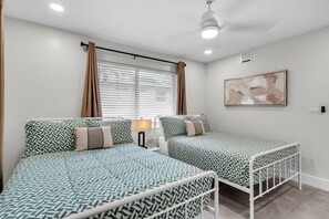 Ein Zimmer mit zwei Betten, einem Nachttisch, einer Lampe, einem Deckenventilator und einem Fenster mit Vorhängen.
