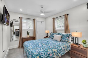 Une chambre à coucher comprenant un lit, des tables de chevet, un miroir, un ventilateur de plafond et une fenêtre avec des rideaux.