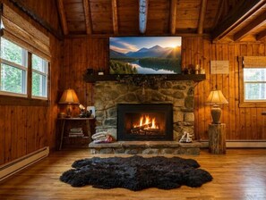 A log cabin interior with a stone fireplace, a television mounted above it, and a large window with a view of mountains and a lake. - Lake Harmony retreat with hot tub, sauna, game room, and ski access (Lake Harmony)