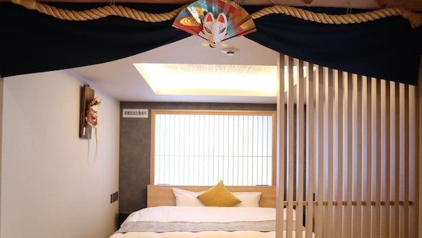A bedroom with a bed, a window with blinds, a ceiling fan, and a decorative mask on the wall. - Kaleido inn Hiroshima (Hiroshima)