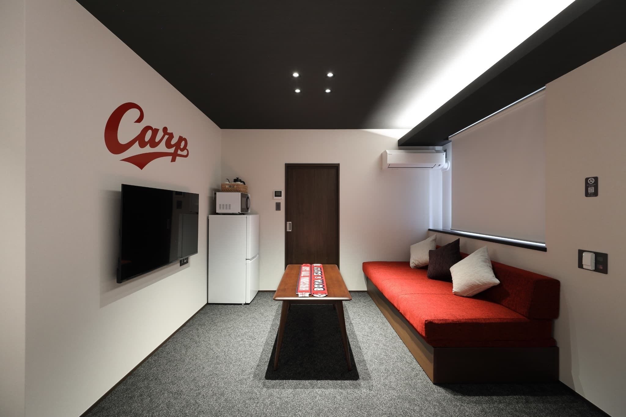 Una sala moderna con un sofá rojo, un televisor de pantalla plana montado en la pared, una mesa de centro y una puerta que da a otra habitación.