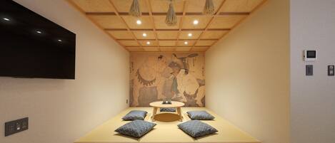 A room with a tatami mat floor, a low table with cushions, a mounted TV, and a traditional Japanese mural on the wall.