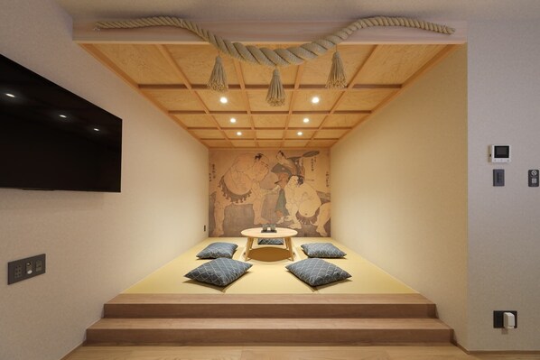 A room with a tatami mat floor, a low table with cushions, a mounted TV, and a traditional Japanese mural on the wall.