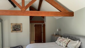 Un dormitorio con cama, mesita de noche, lámpara, sofá gris, techo de madera, puerta y un cuadro enmarcado en la pared.