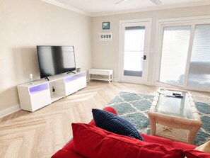 A living room with a flat-screen TV on a white cabinet, a red sofa, a glass coffee table, and a wicker chair. - Peaceful 2-bedroom condo with AC in Panama City Beach (Panama City Beach)