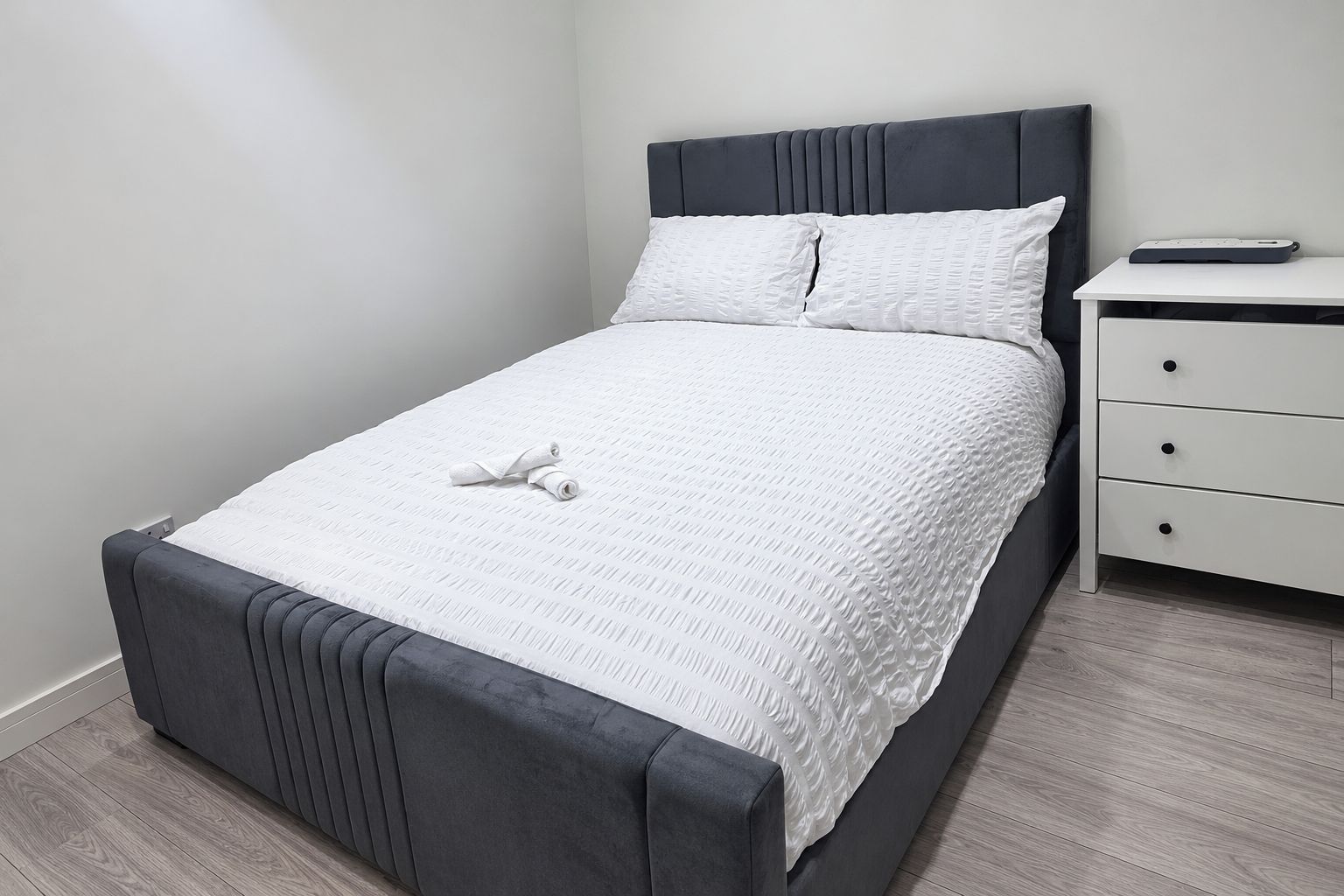 A neatly made bed with a grey headboard and white bedding, a bedside table with a book, and a drawer unit.