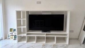 Ein modernes Wohnzimmer mit einem Flachbildfernseher auf einer weißen Unterhaltungseinheit, einem Fliesenboden und einer kleinen Kunstausstellung links.