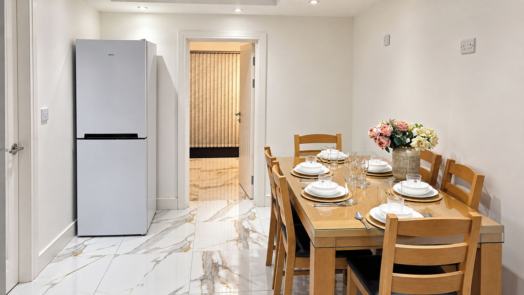 A dining area with a table set for four, a refrigerator, and a door leading to a room with blinds.