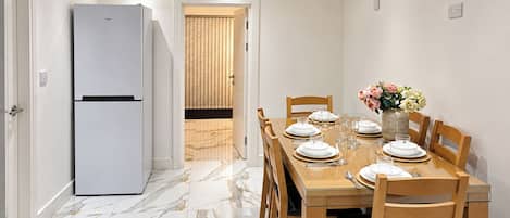 A dining area with a table set for four, a refrigerator, and a door leading to a room with blinds.