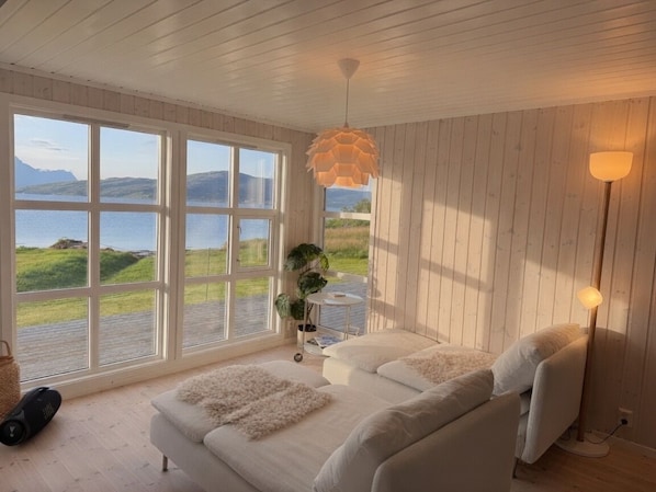 A room with a large window offering a view of a lake and hills, a white sofa with fluffy cushions, a hanging light fixture, and a floor lamp.
