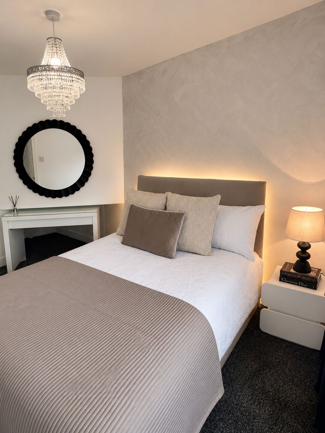 A hotel room with a large bed, a bedside table with a lamp, a round mirror, and a chandelier.