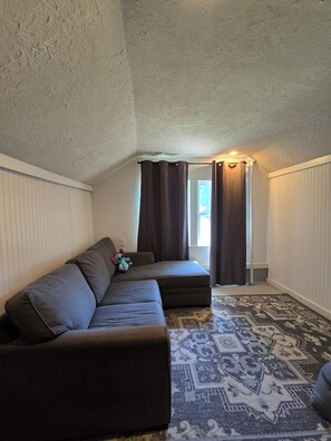 Ein Zimmer mit Ecksofa, einem gemusterten Teppich und einem Fenster mit Vorhängen.