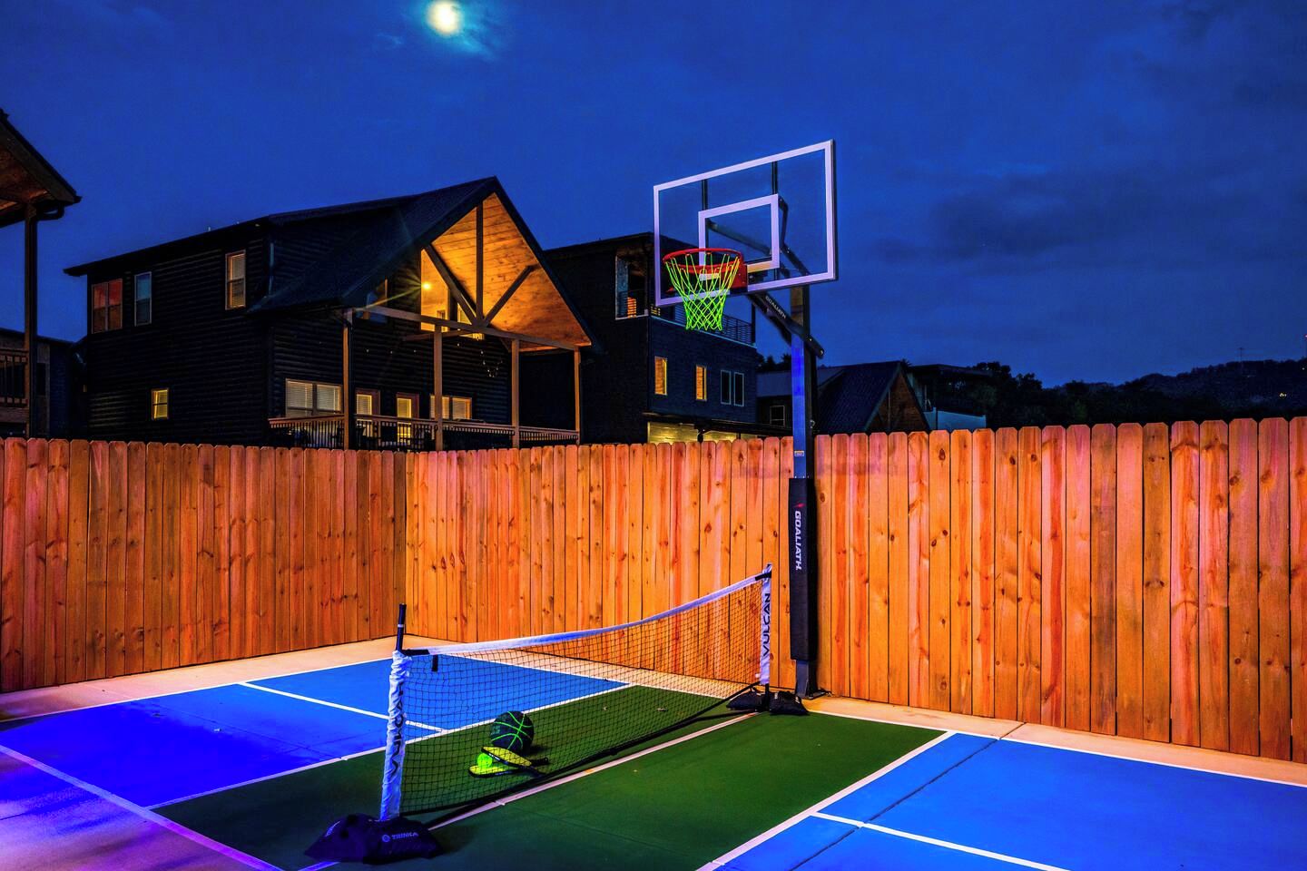 Un terrain de basket-ball en plein air, entouré d’une clôture en bois, avec un panier de basket et un filet de tennis.