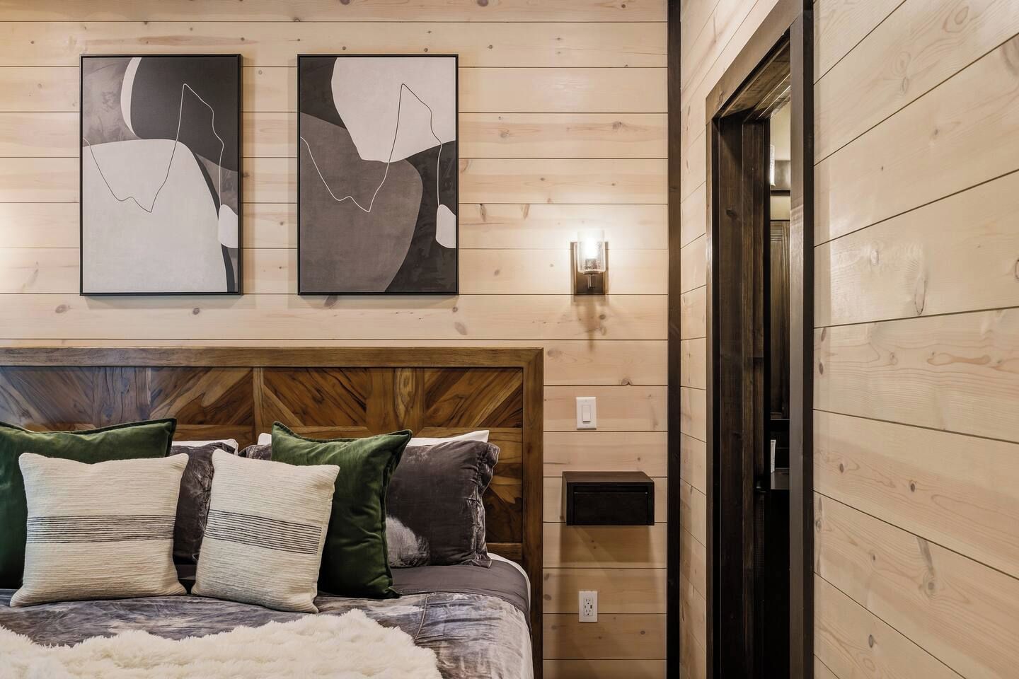Quarto com cabeceira de madeira, cama com várias almofadas e duas pinturas abstratas na parede.