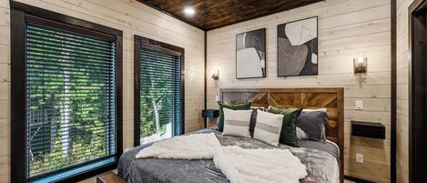 A bedroom with a large bed, wooden walls, and a view of greenery through the windows.