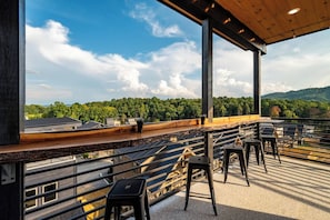 Eine Dachterrasse mit Blick auf eine bewaldete Landschaft, mit einer Holzterrasse, Metallgeländern und schwarzen Barhocker.