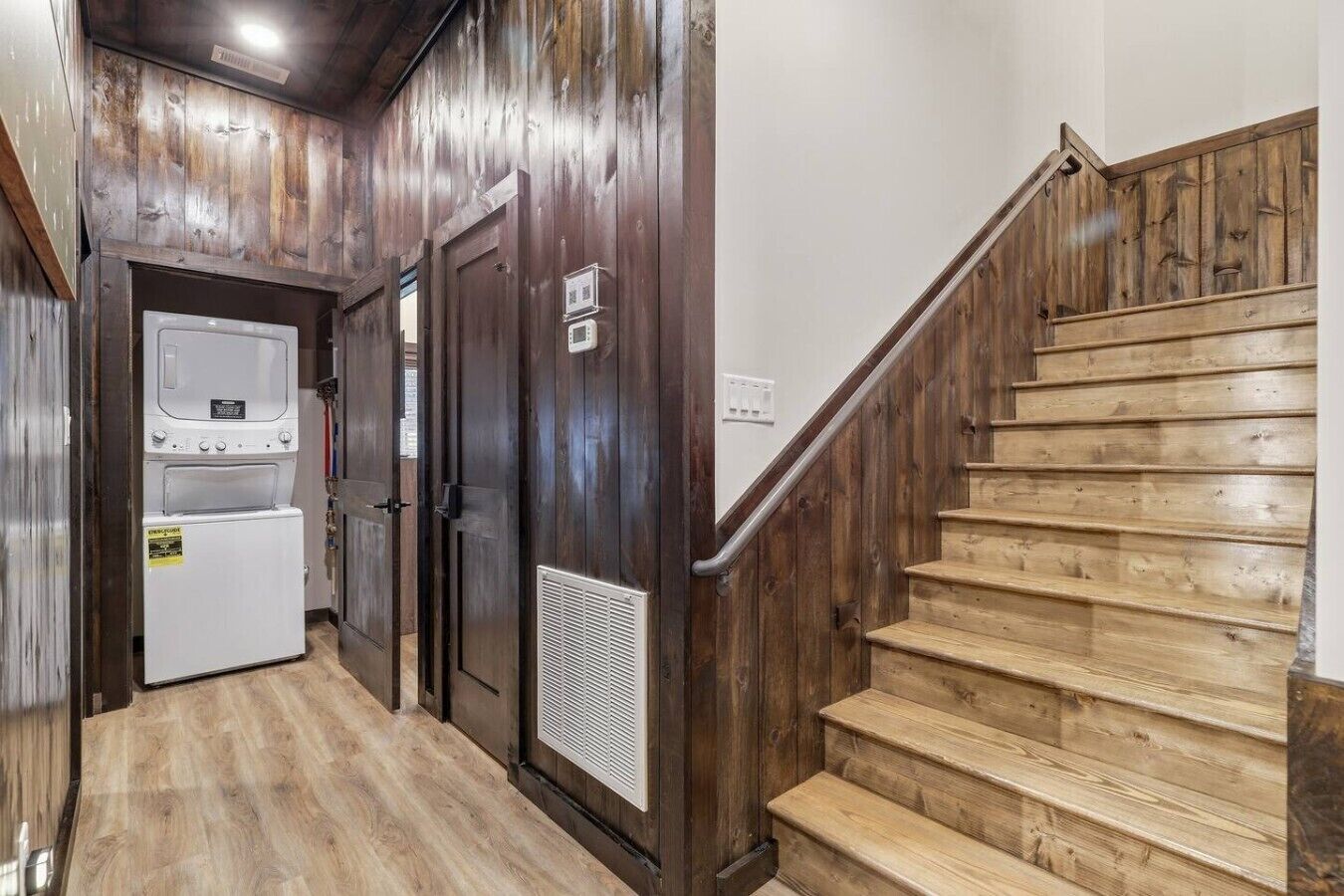 A wooden staircase leading to a landing with a laundry room featuring two dryers and a vent.