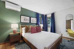 Una habitación de hotel con cama, mesitas de noche, lámpara, una silla, ventana con cortinas y una obra de arte en la pared.
