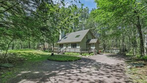 Une cabane dotée d’un porche, entourée d’arbres, et une allée de gravier menant à l’entrée.
