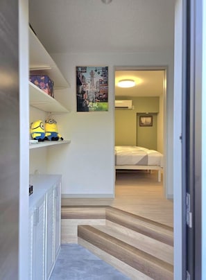 Een gang met een bed op de achtergrond, een plank met spullen en een schilderij aan de muur.