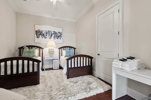A child's bedroom with two cribs, a bedside table with a lamp, a white door, and a framed picture on the wall.