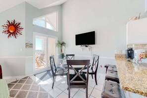 A modern kitchen with a flat-screen TV, a dining area with chairs, and a sun decoration on the wall. - Beachfront Gulf Shores condo with ocean view, pool, hot tub & balcony. (Gulf Shores)