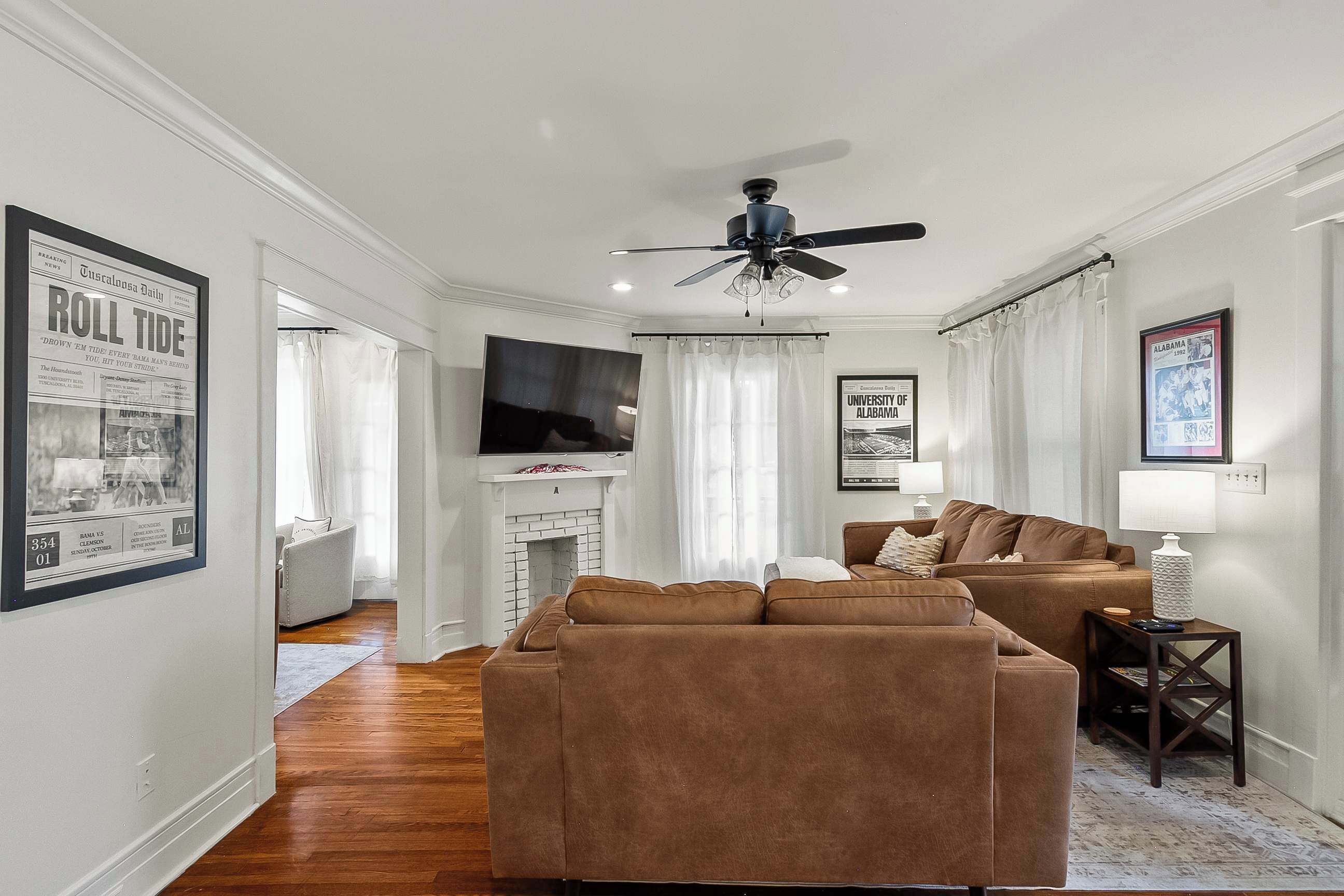 A living room with a brown sofa, a fireplace, a ceiling fan, and framed pictures on the walls.