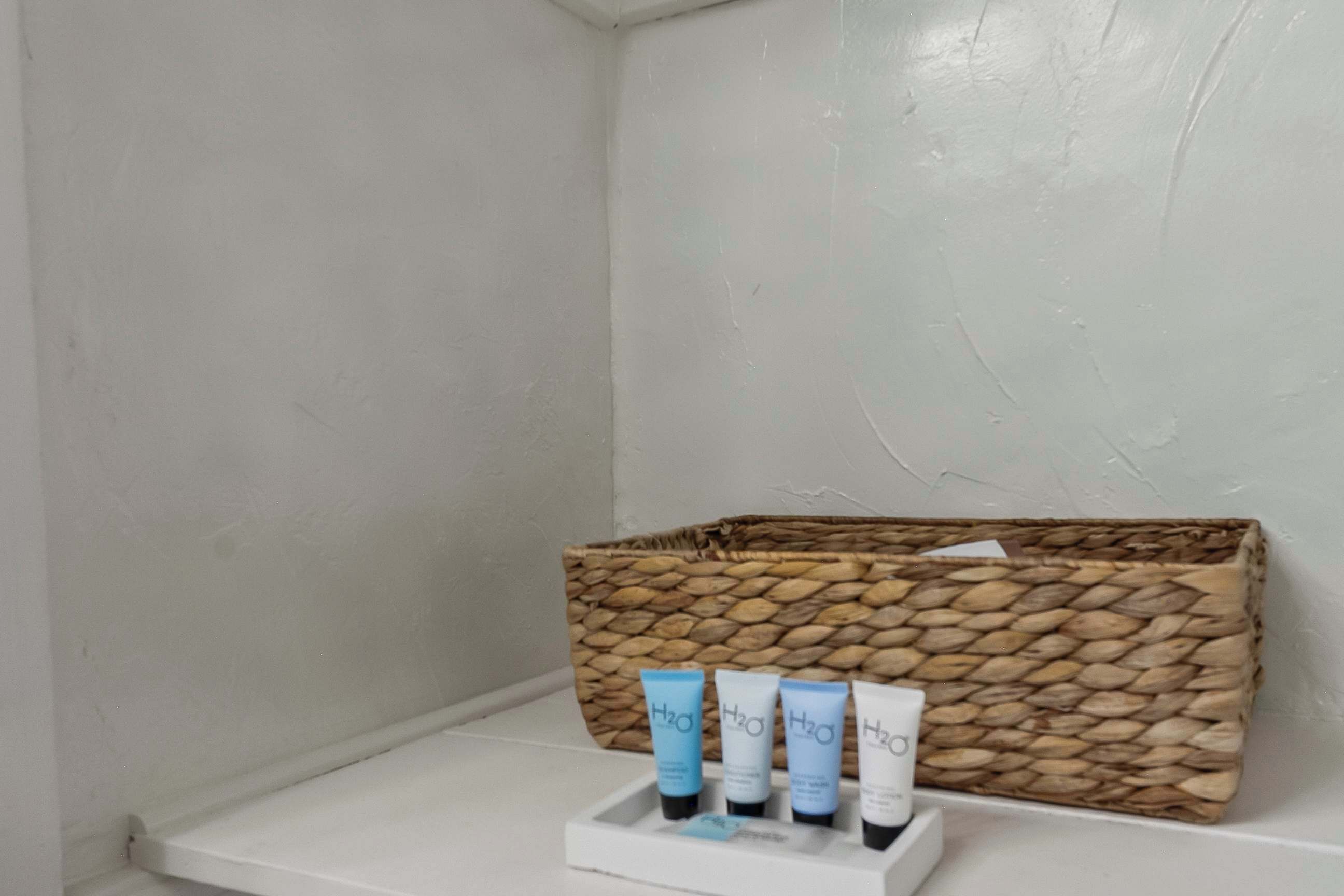 Un baño con un lavamanos blanco, un estante blanco y una cesta tejida con cuatro botellas de H2O.