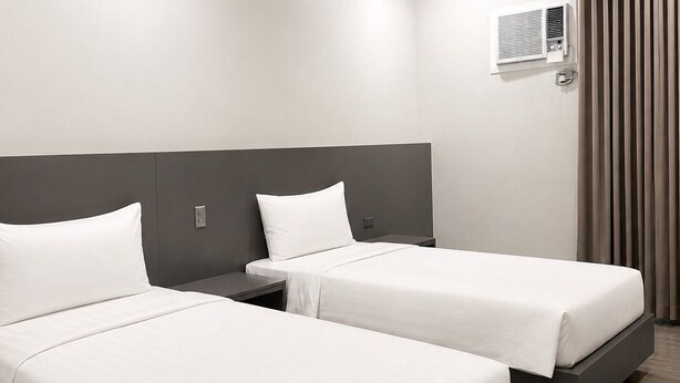 A hotel room with two beds, a ceiling-mounted air conditioning unit, and a window with curtains.