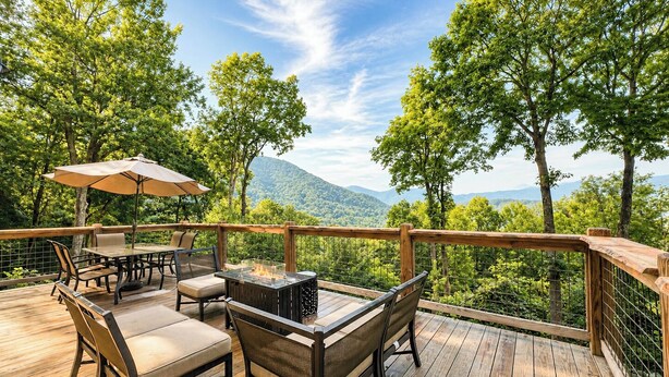 An outdoor deck with a table and chairs, surrounded by a wooden railing and overlooking a forested landscape. - Long-Range Views on 3 Levels | Hot Tub + Fire Pit (Maggie Valley)