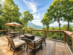 An outdoor deck with a table and chairs, surrounded by a wooden railing and overlooking a forested landscape. - Long-Range Views on 3 Levels | Hot Tub + Fire Pit (Maggie Valley)