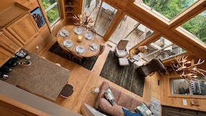 A wooden cabin interior with a dining area, seating, and a large window overlooking greenery. - Long-Range Views on 3 Levels | Hot Tub + Fire Pit (Maggie Valley)
