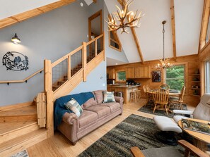 A spacious loft with a wooden staircase, a cozy living area with a sofa and chairs, a dining table, and a kitchen with wooden cabinets. - Long-Range Views on 3 Levels | Hot Tub + Fire Pit (Maggie Valley)