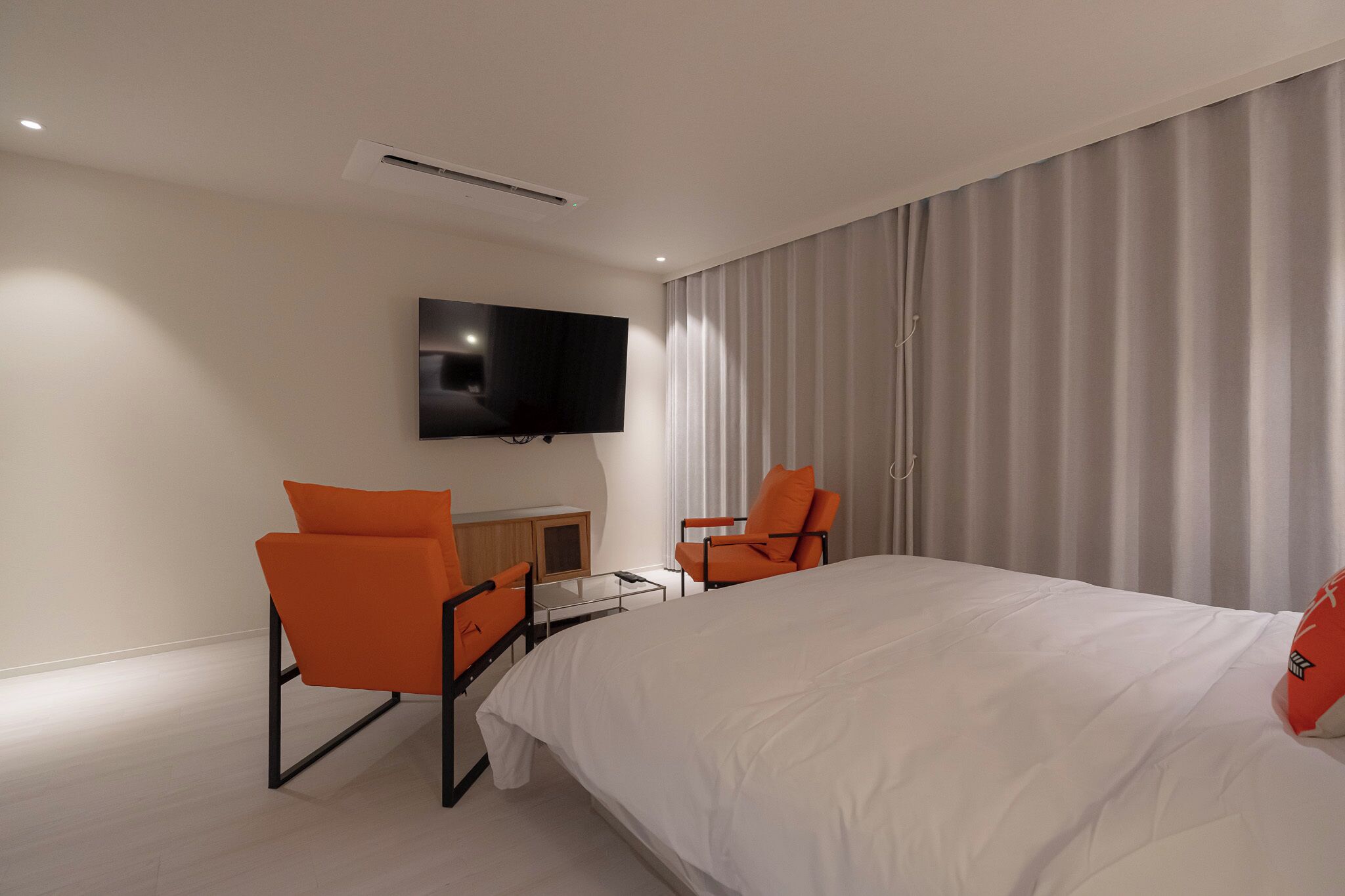 Ein modernes Hotelzimmer mit einem großen Bett, zwei orangenen Sesseln, einem flachbildfernseher an der Wand und einem großen Fenster mit Vorhängen.