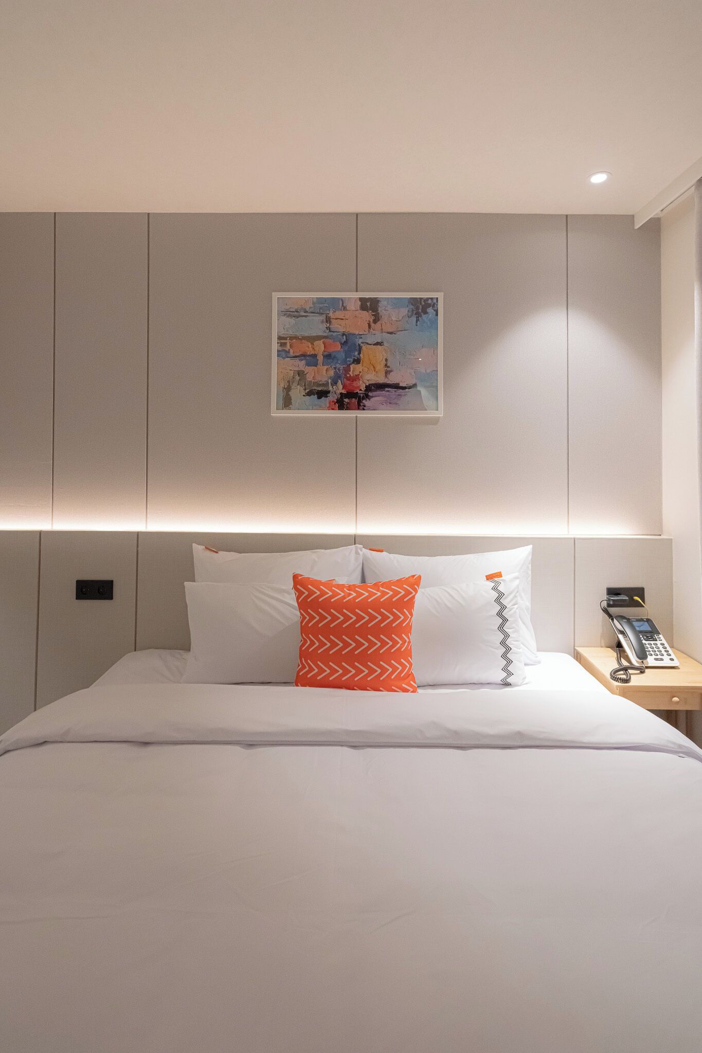 A hotel room with a bed, pillows, a bedside table with a phone, and a painting on the wall.