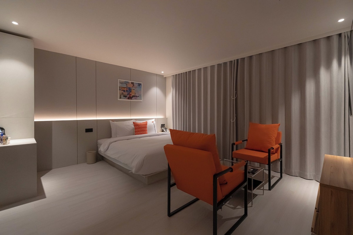 A modern hotel room with a bed, two orange chairs, a bedside table, and a large window with curtains.