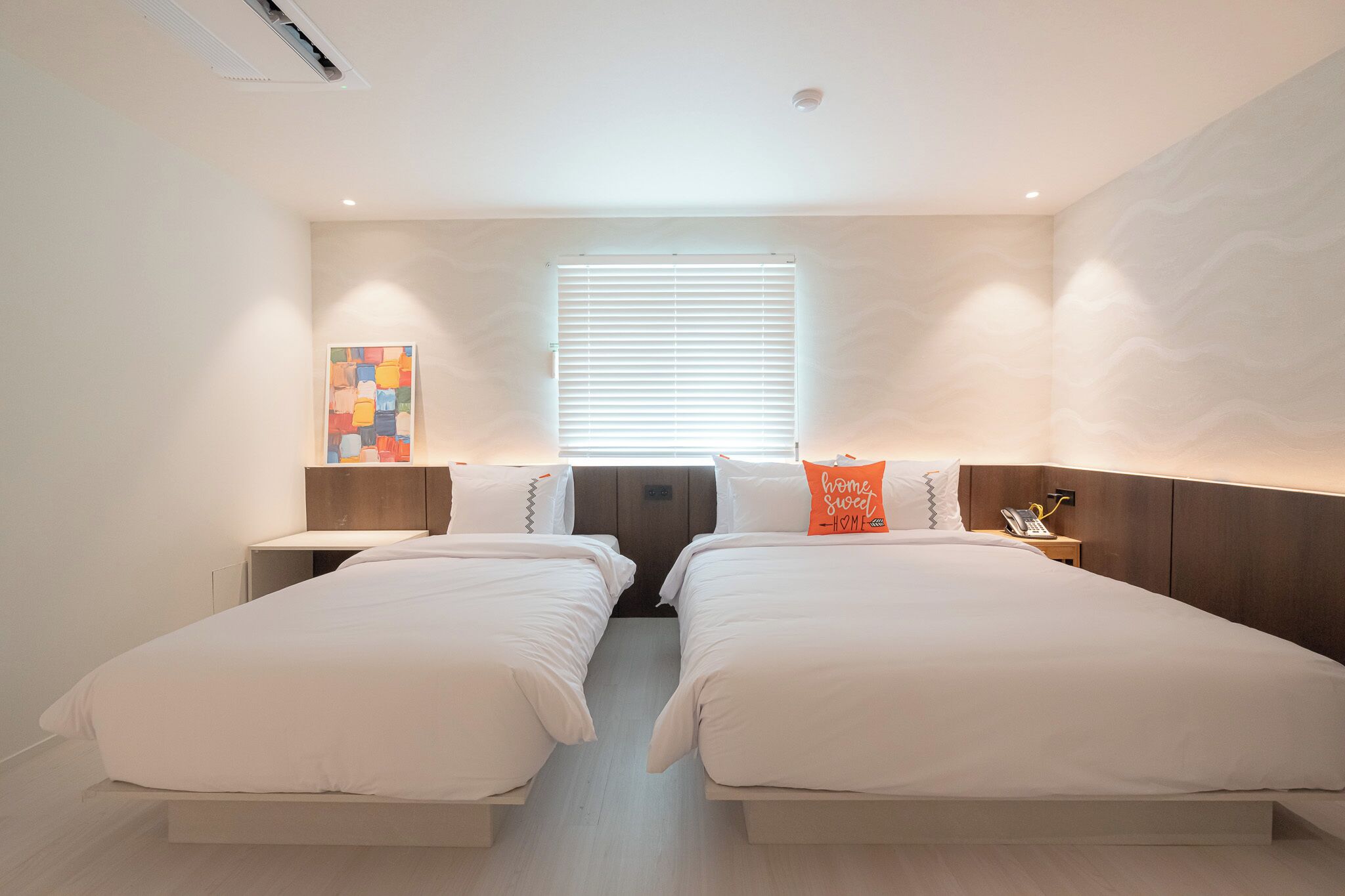 Habitación doble con dos camas, una ventana con persianas, una mesita de noche con lámpara y una obra de arte en la pared.