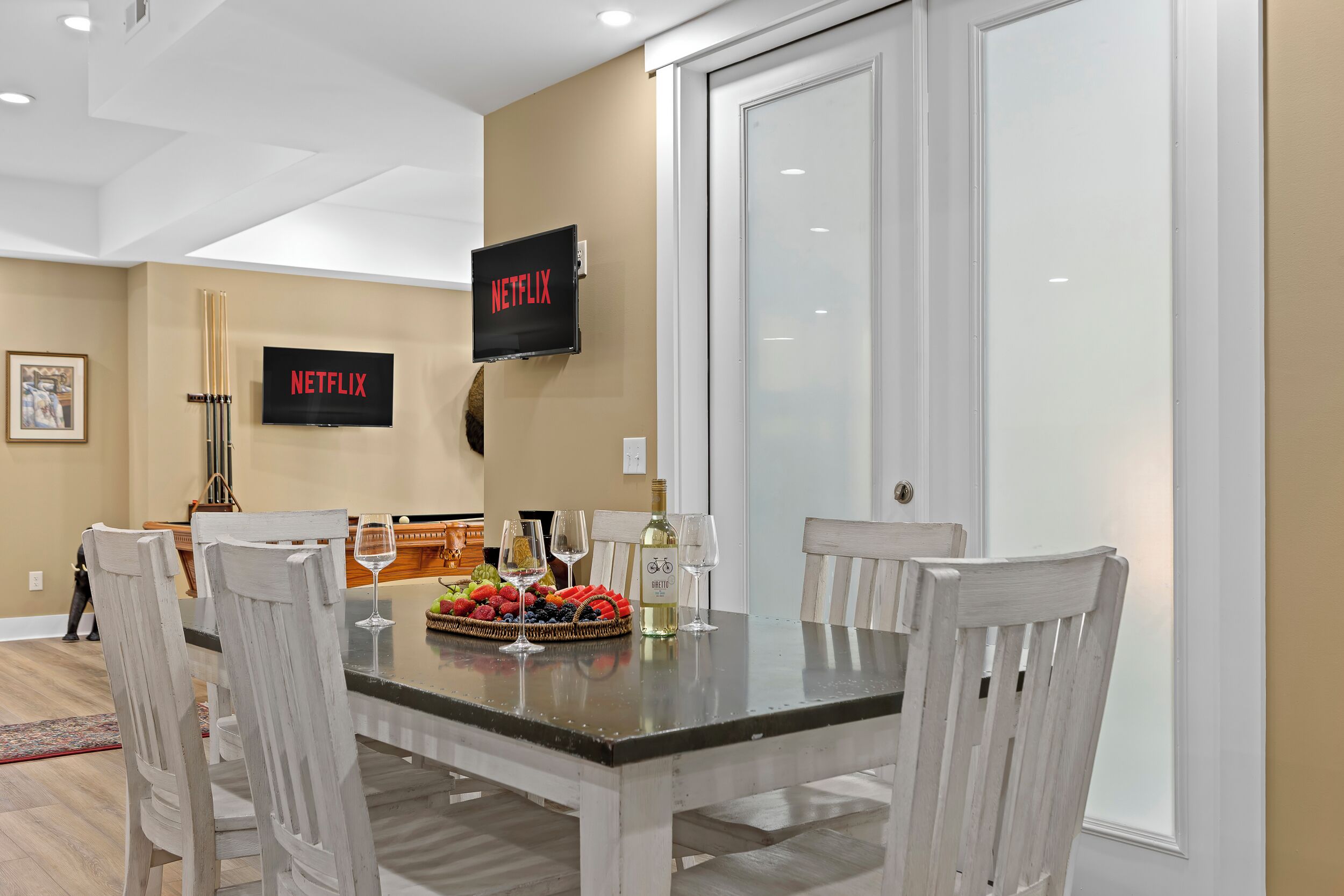 A dining area with a glass table, white chairs, and a wall-mounted TV displaying the Netflix logo.