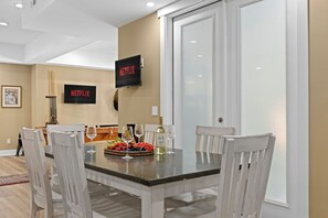 A dining area with a glass table, white chairs, and a wall-mounted TV displaying the Netflix logo.