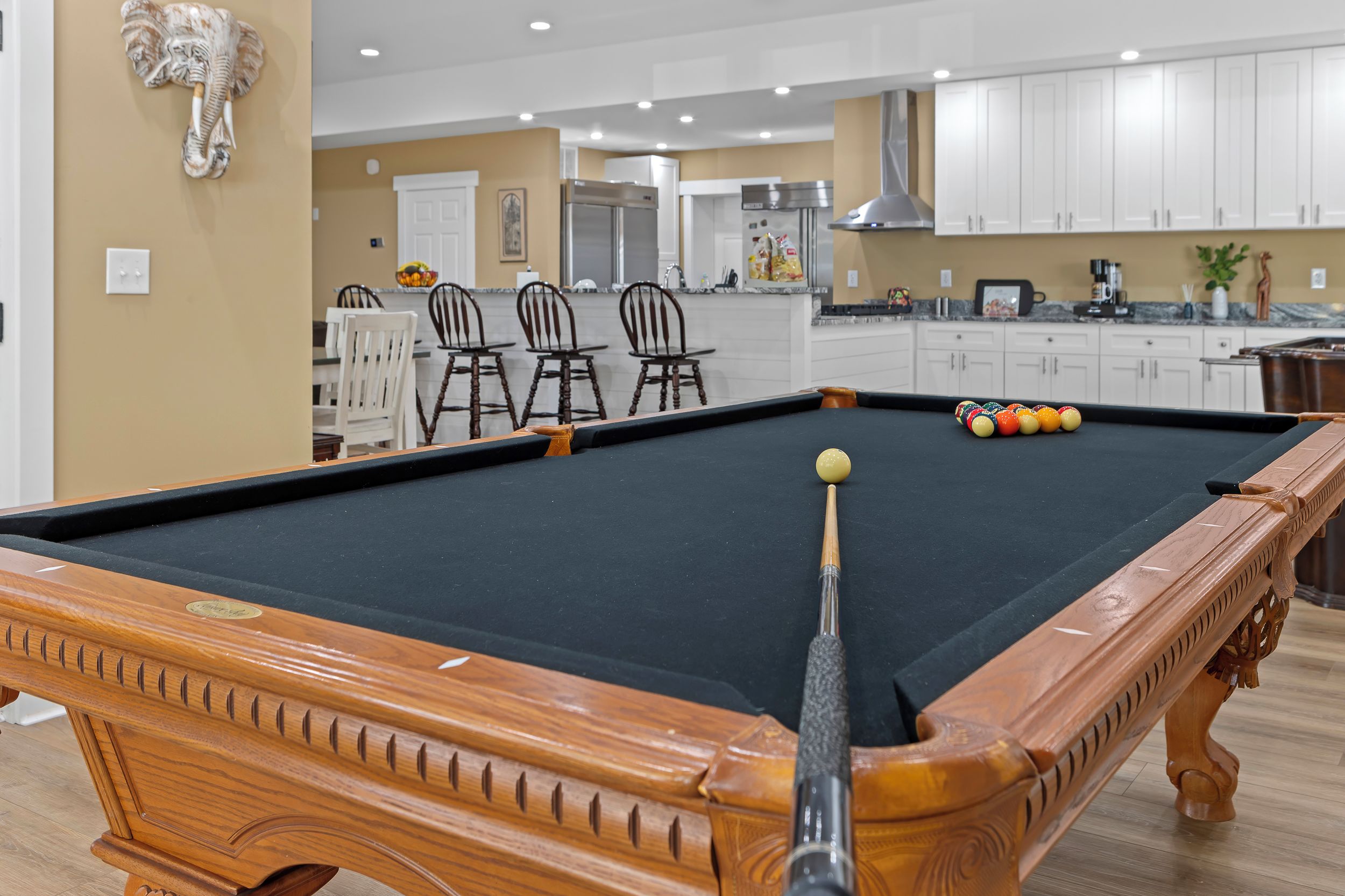 A pool table with a cue stick, a dining area with chairs, and a kitchen in the background.