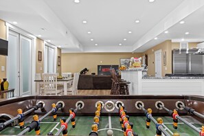 A modern living room with a foosball table, a dining area, and a kitchen with a refrigerator.
