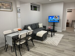 A modern living room with a grey sofa, a dining table with chairs, a flat-screen TV, and a small side table. - Comfortable modern basement suite (Winnipeg)
