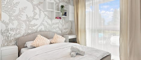 A bedroom with a bed, nightstands, a wall-mounted shelf, and a large window with curtains.