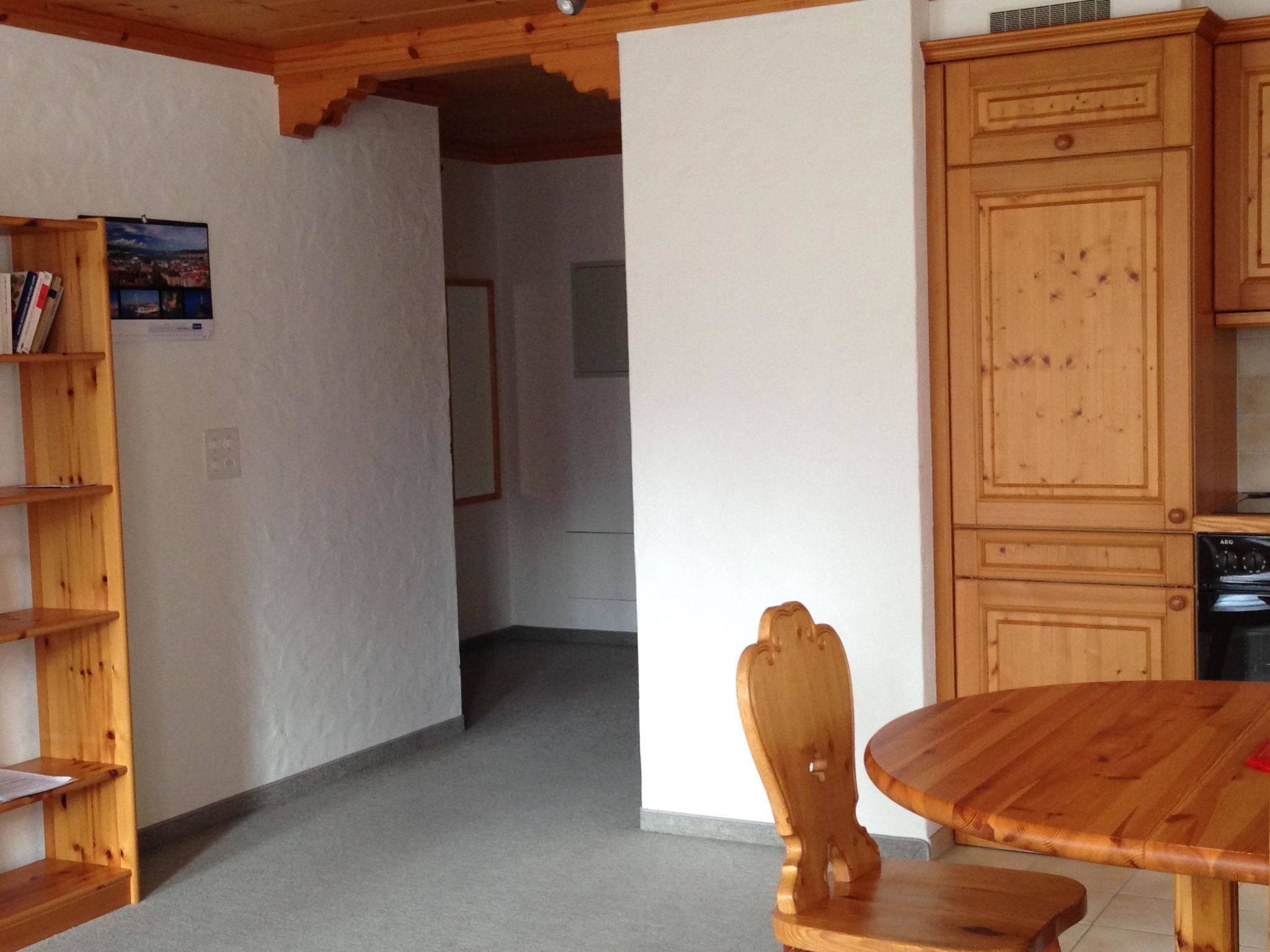 Een houten eettafel met een stoel, een houten kast en een boekenkast in een kamer met een vloerkleed en een witte muur.