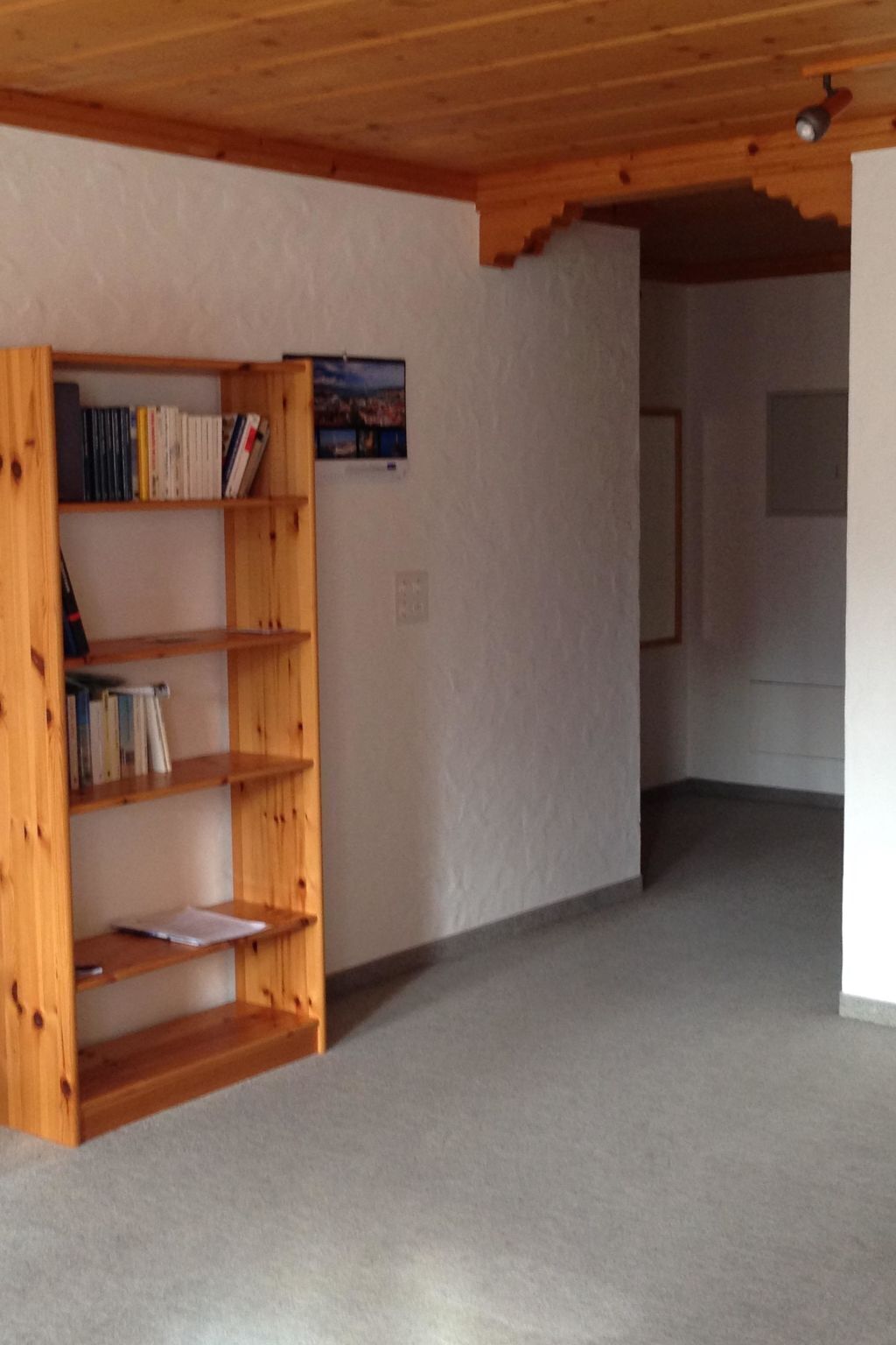A room with a wooden bookshelf against a wall, a carpeted floor, and a ceiling with wooden beams.
