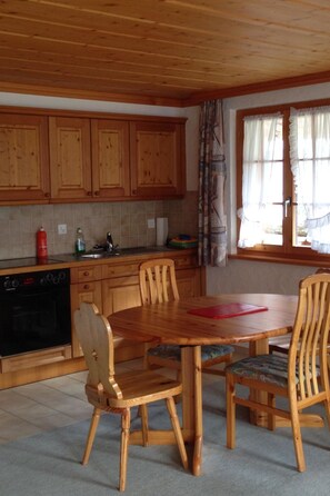 A kitchen with wooden cabinets, a dining table with chairs, a red cutting board, and a window with curtains. - Favre OG Ost Appartement/Fewo, Dusche oder Bad, WC, 1 Schlafraum 2-Bett-Wohnung by Interhome (Matten  (St. Stephan))